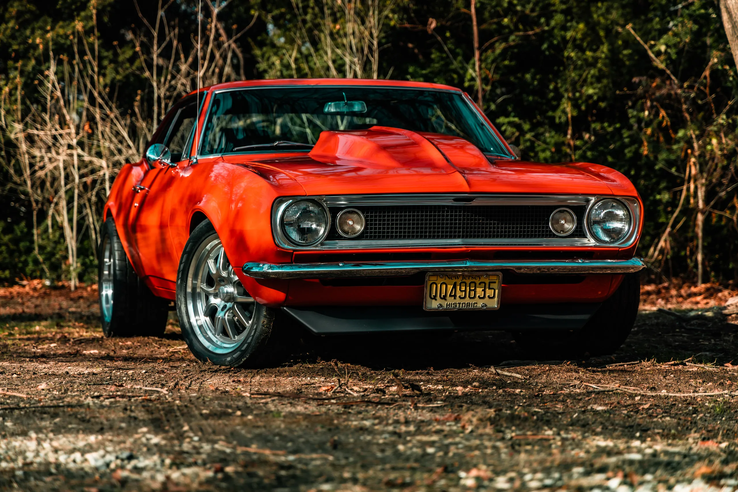 A red first-generation Camaro sits front-three-quarter on a leaf-covered clearing