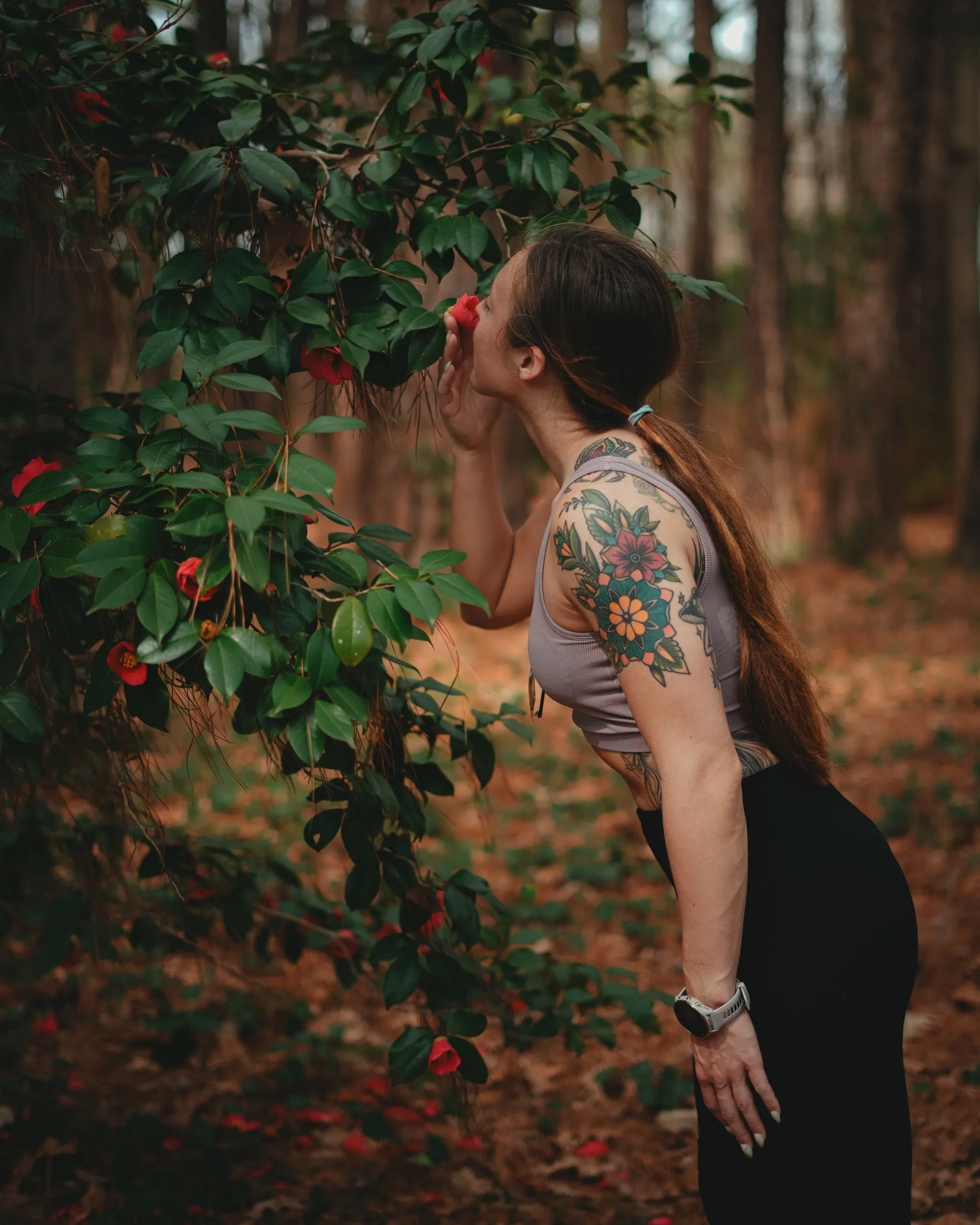 A tattooed woman reaches up to a flowering camellia branch in autumn woods