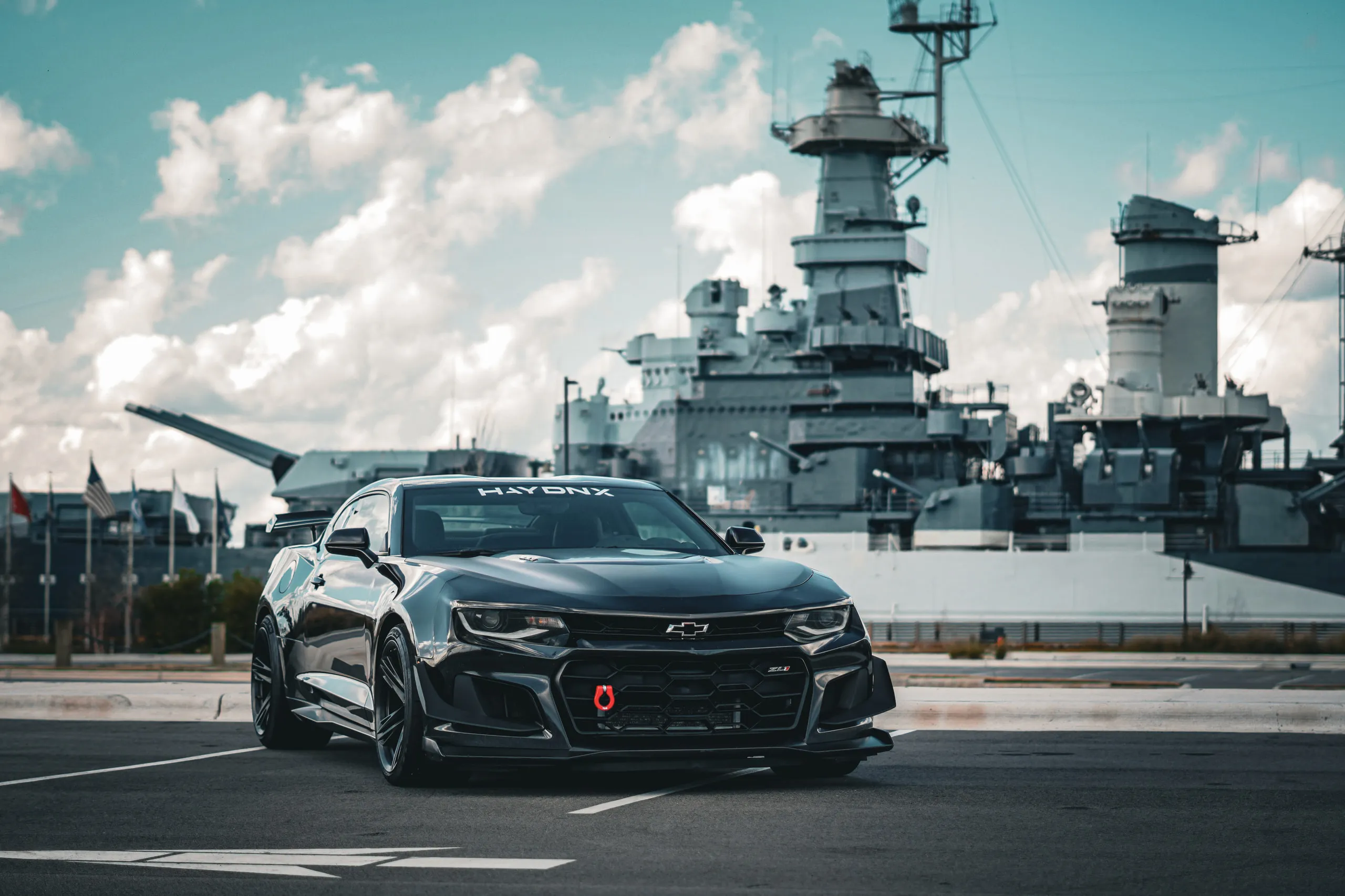 A black Camaro parked on the waterfront in front of a moored battleship