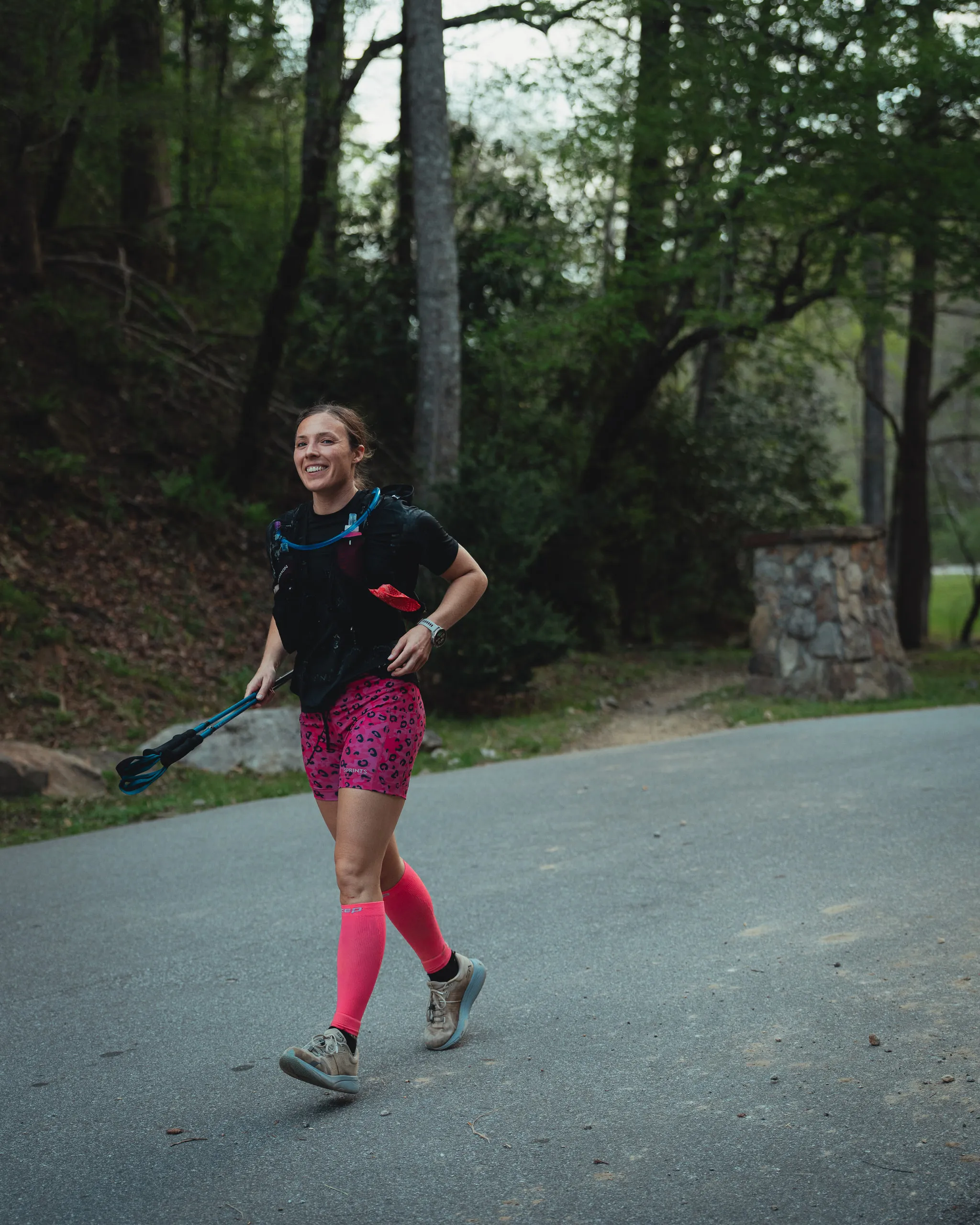 A competitor in pink compression socks runs a paved stretch carrying a weight at her side