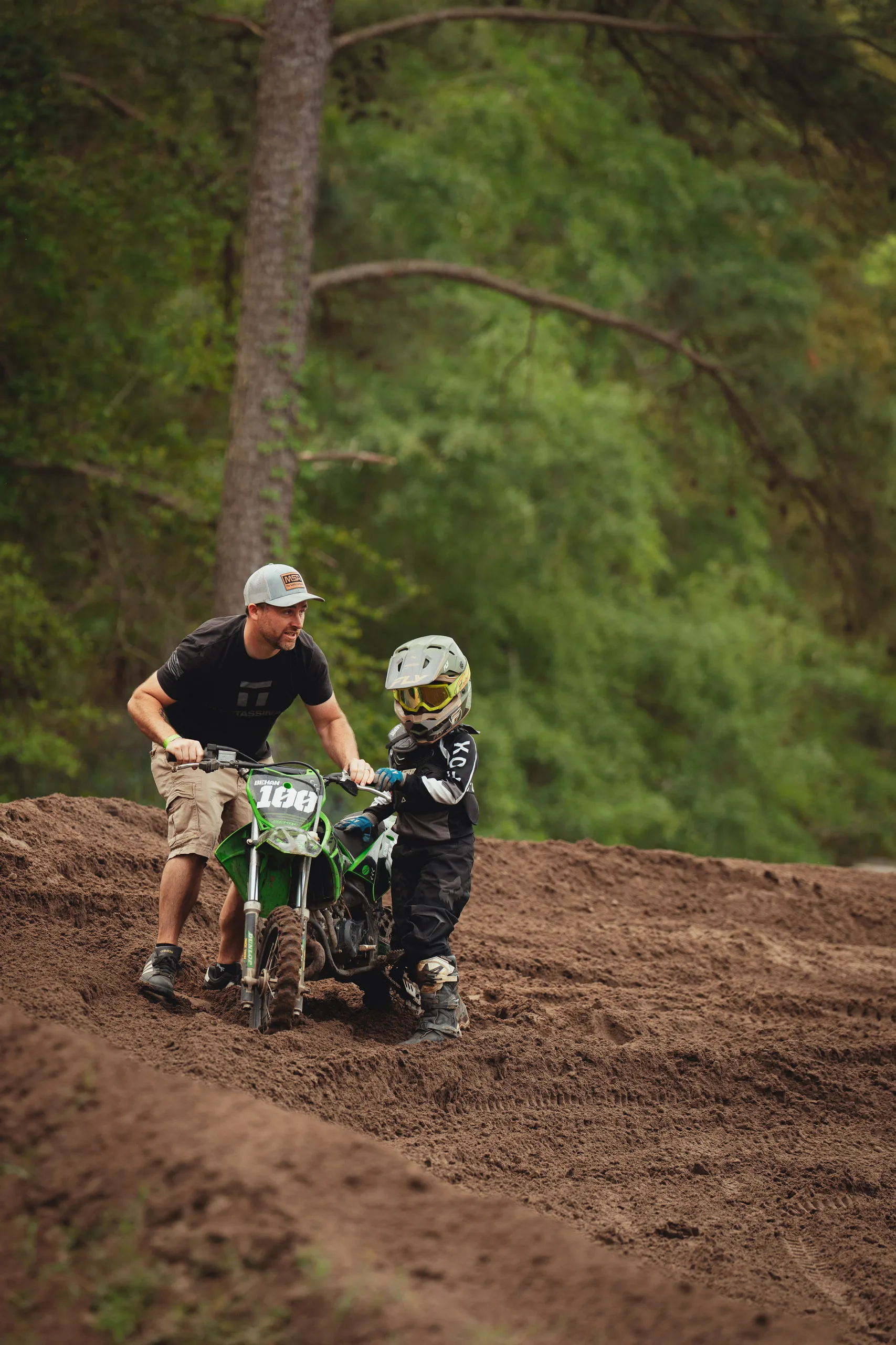 An adult steadies a young rider on a green dirt bike at the top of a rutted climb