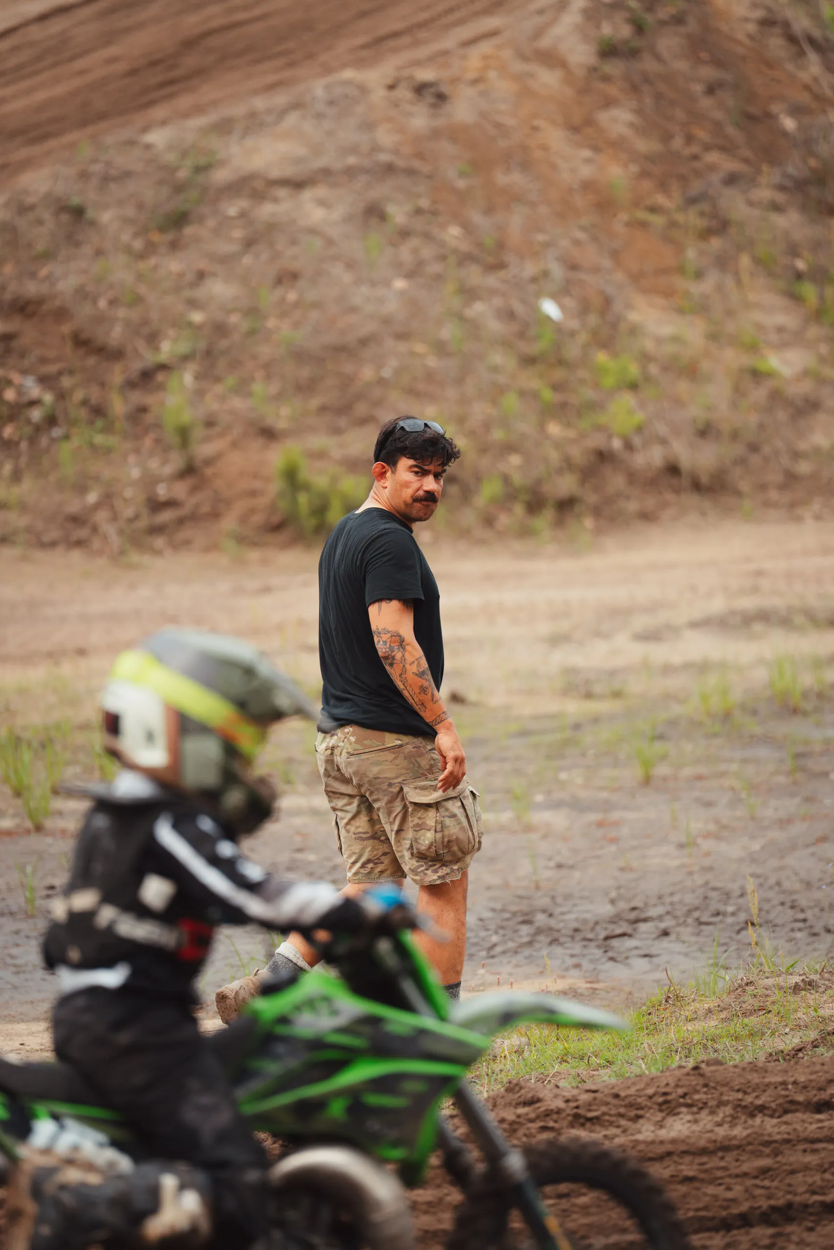 A helmeted child on a mini bike waits while a man in a black shirt watches from the track edge