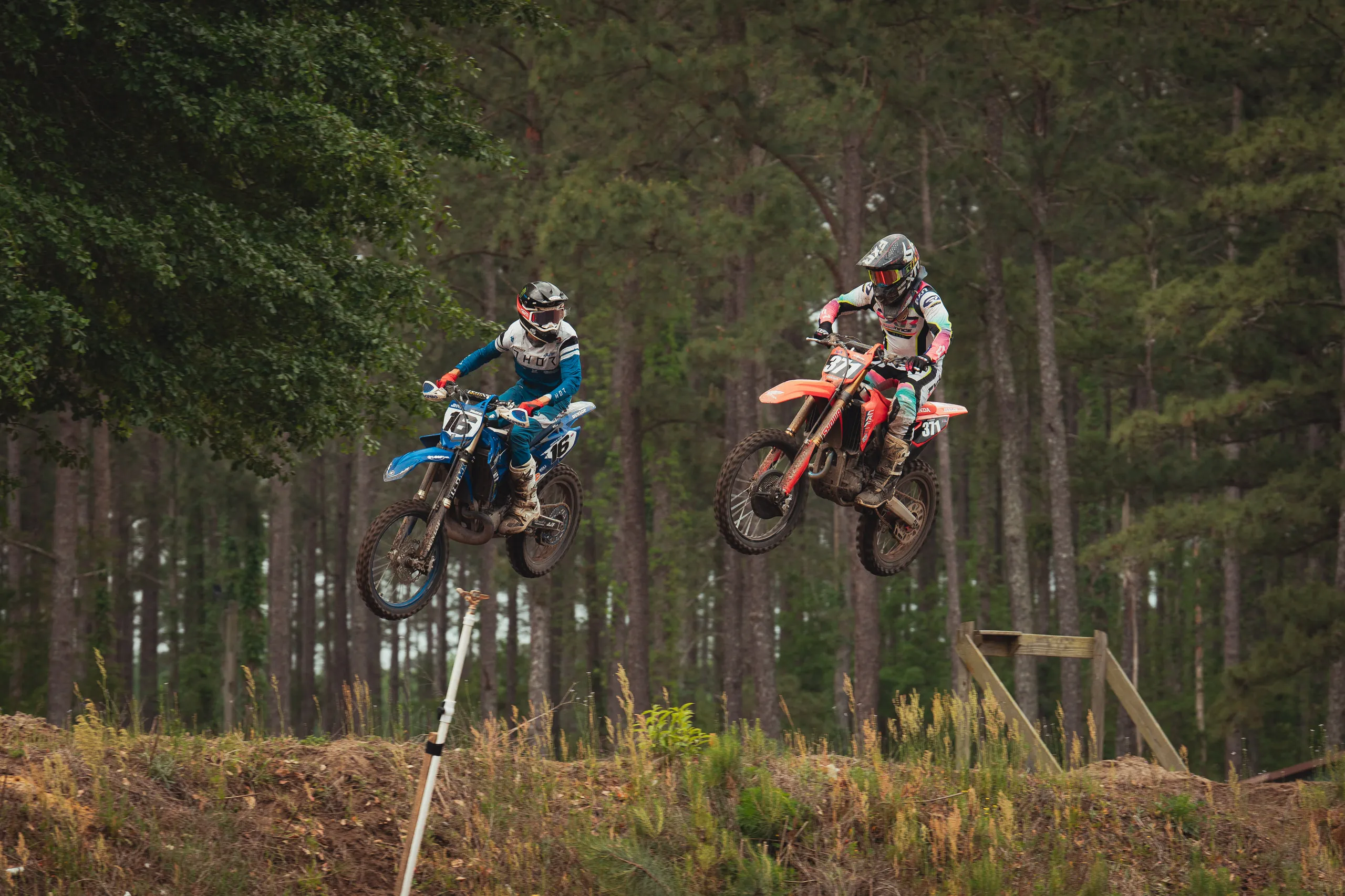 Two riders leap a jump side by side, suspended above the track
