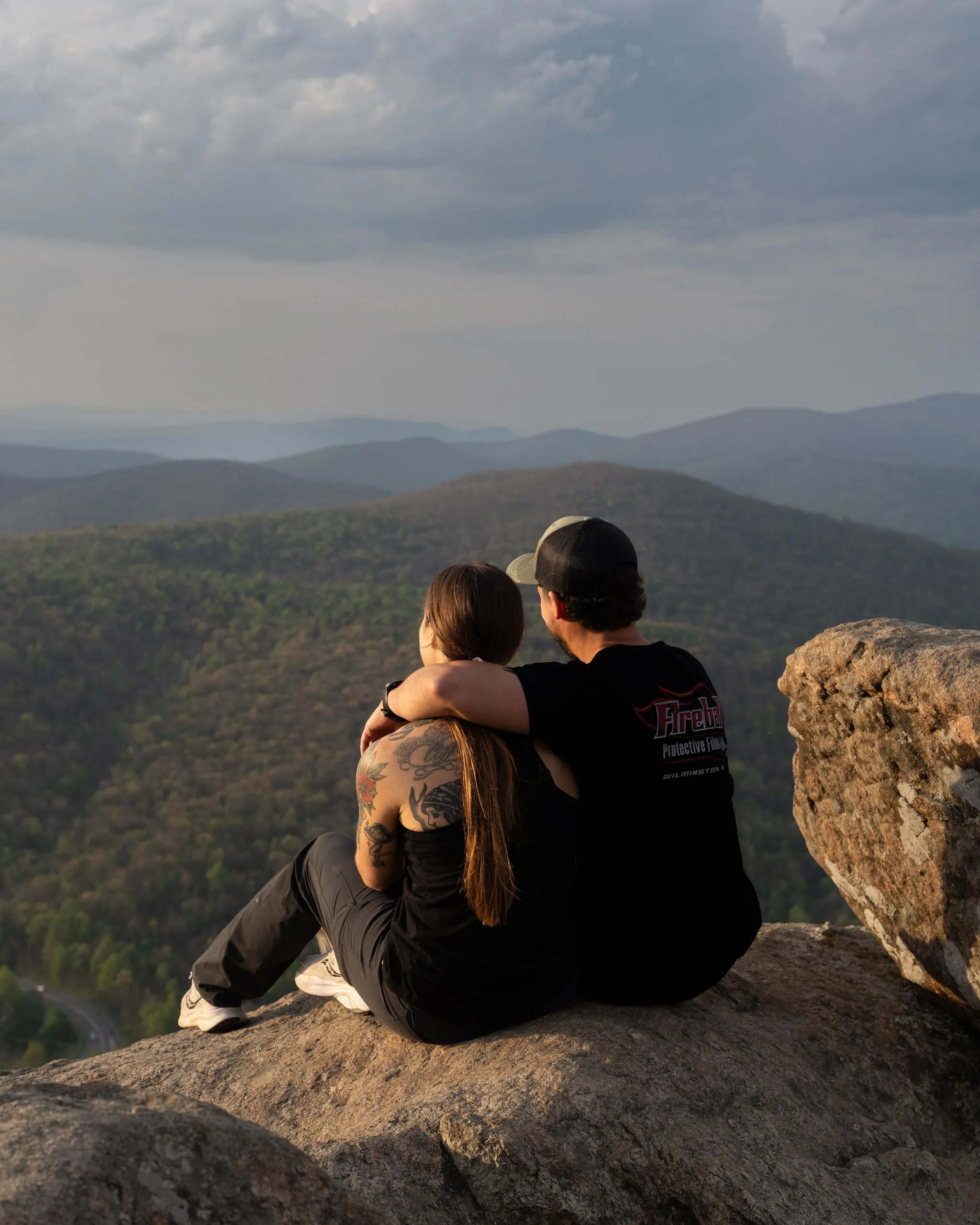A couple sit together on a rock outcrop at golden hour holding a small child, mountains beyond