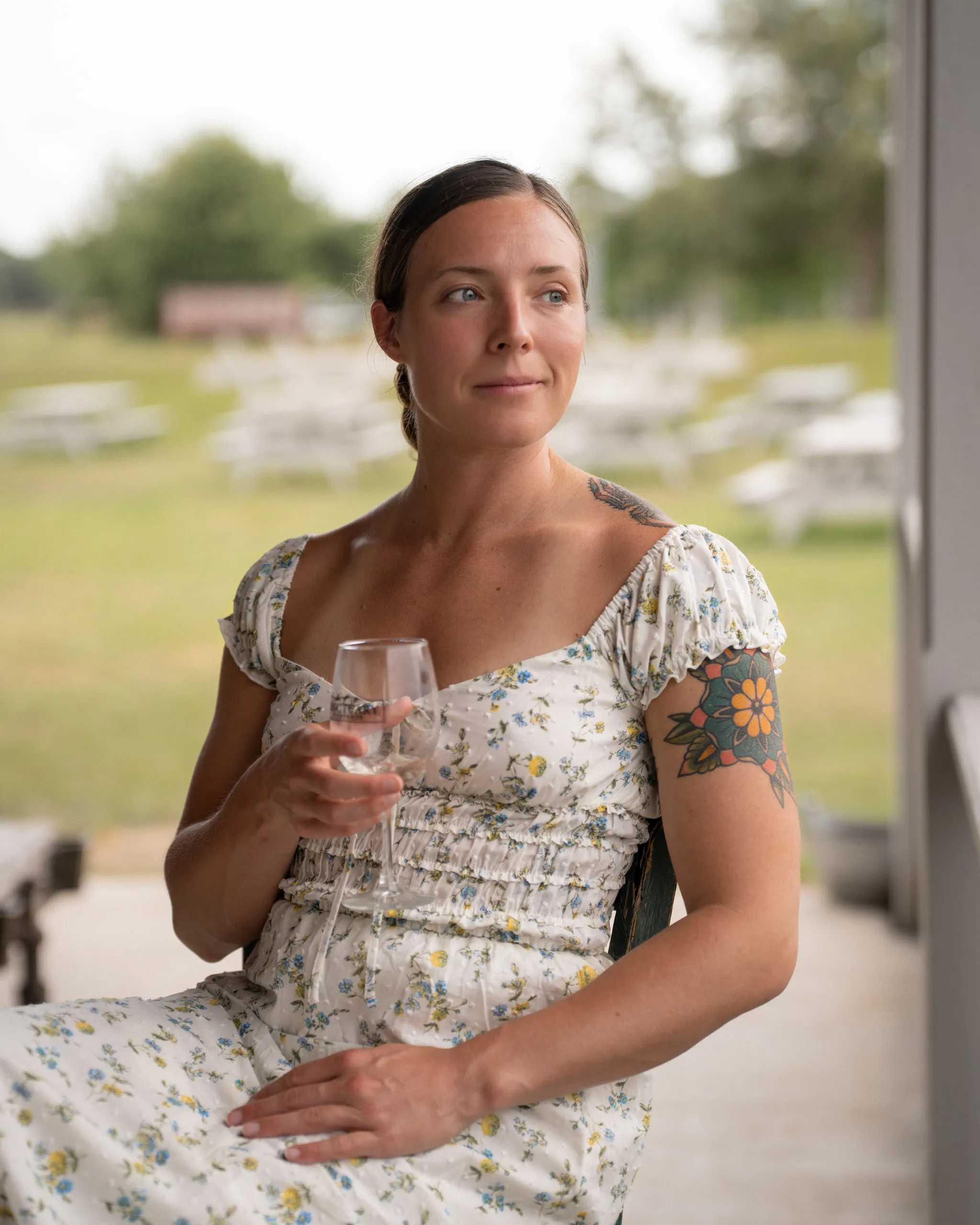 A woman in a floral dress holds a glass of wine in a doorway, orchard behind her