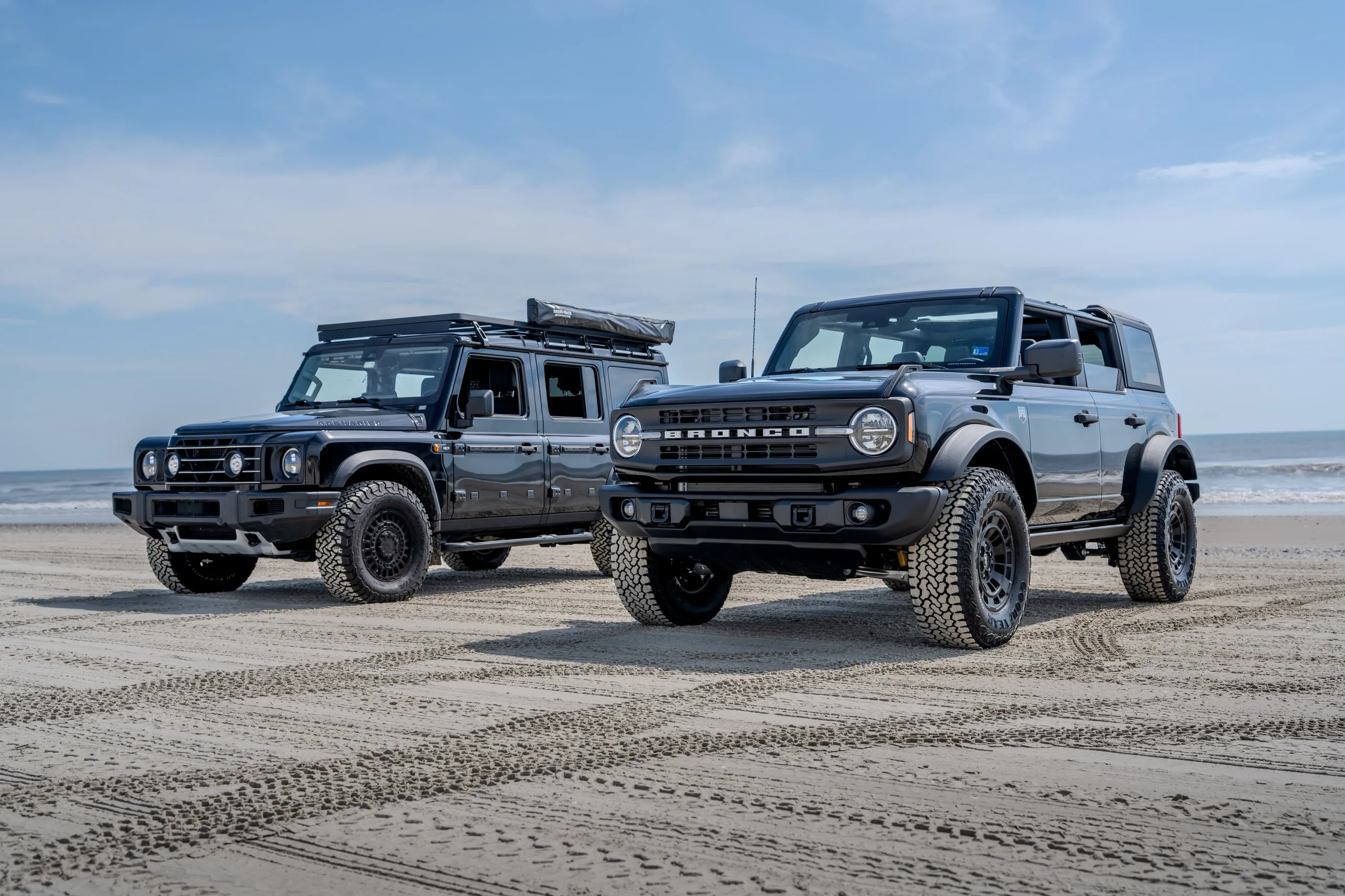 A Ford Bronco and a Grenadier parked side by side on hard-packed sand at the waterline