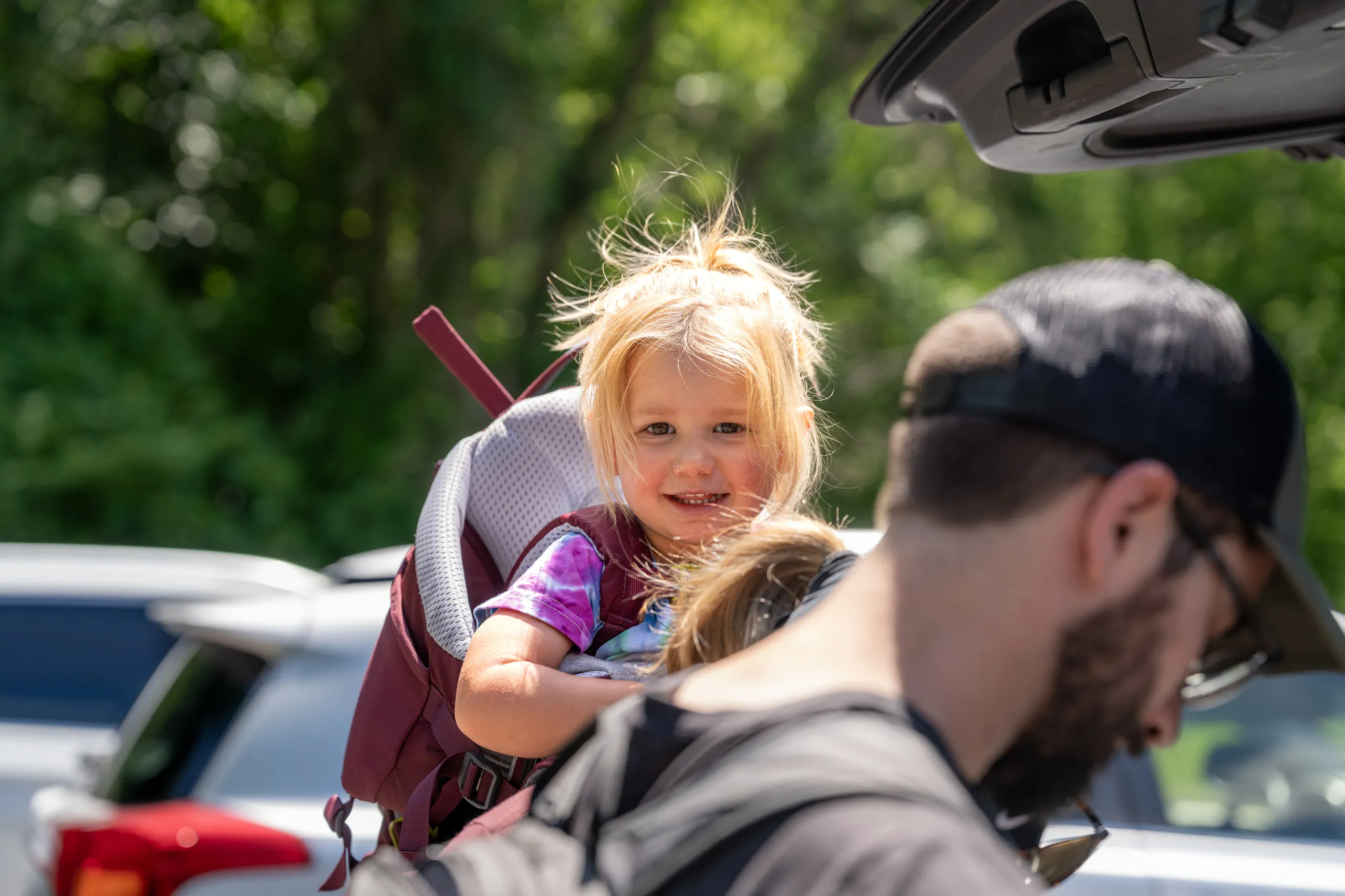 A toddler grins from a backpack carrier over her father's shoulder in a parking lot