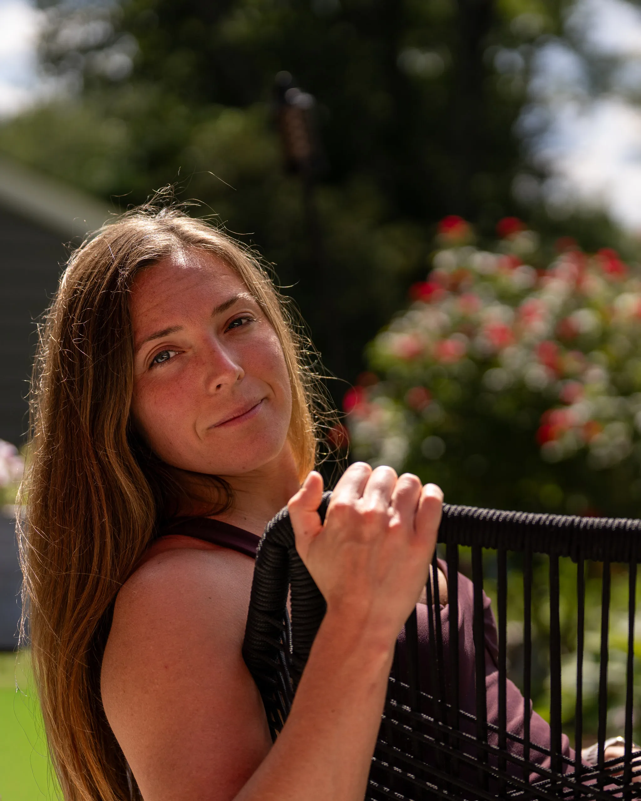 A woman leans on a railing and smiles, flowering shrubs blurred behind her