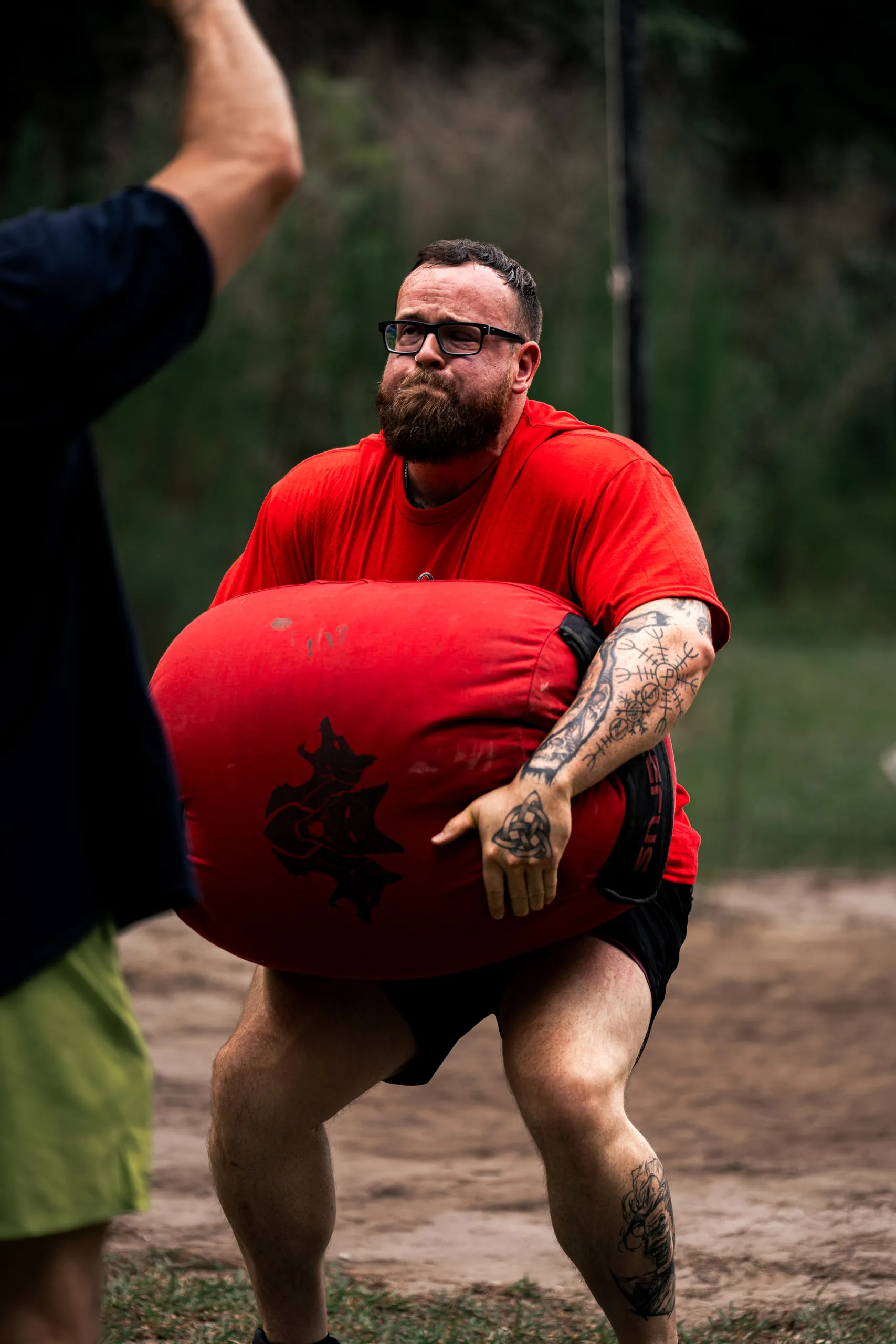 A bearded competitor hauls a heavy red sandbag across the grass, arms locked underneath