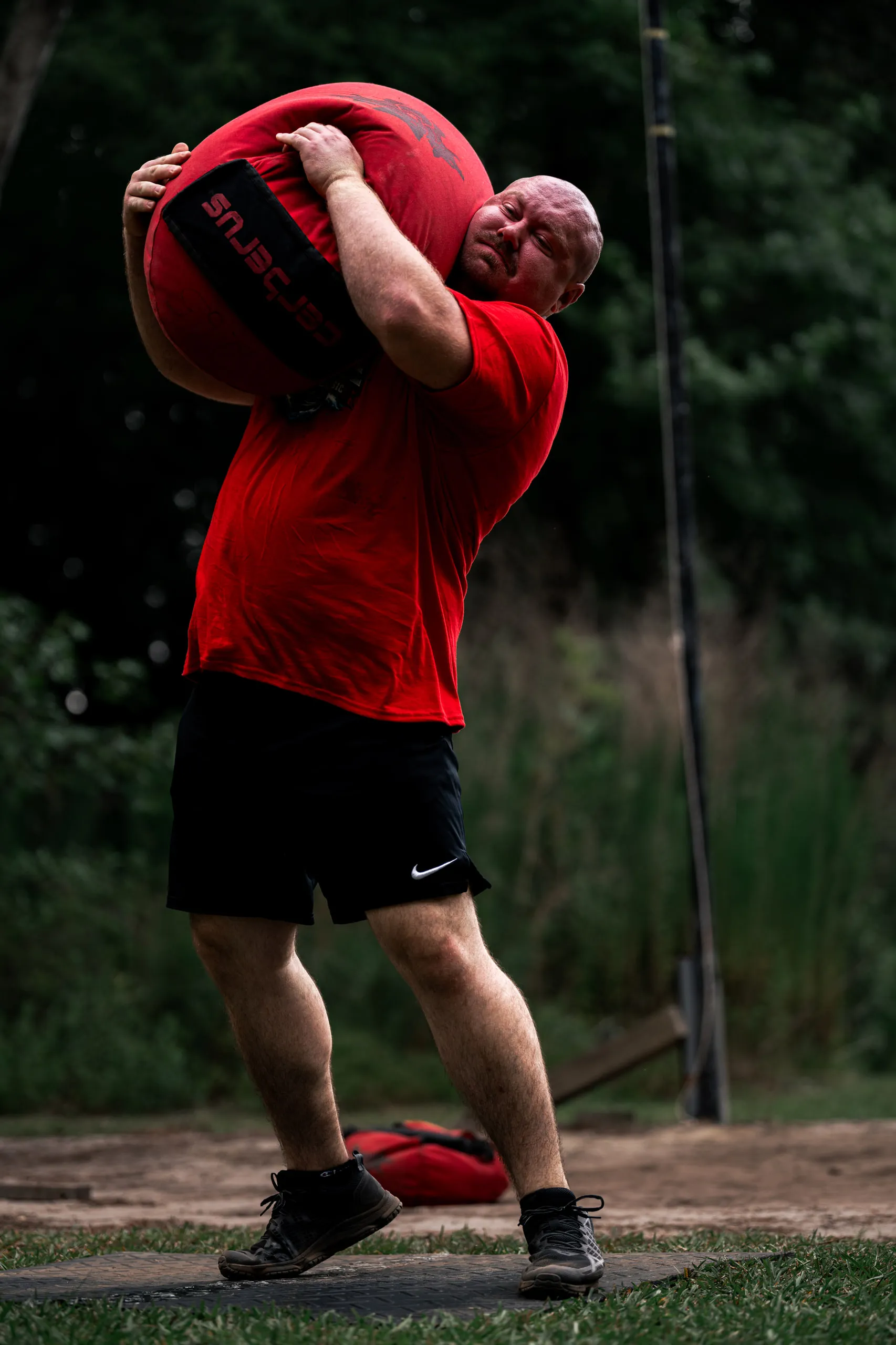A competitor shoulders a heavy red sandbag, head turned up under the load