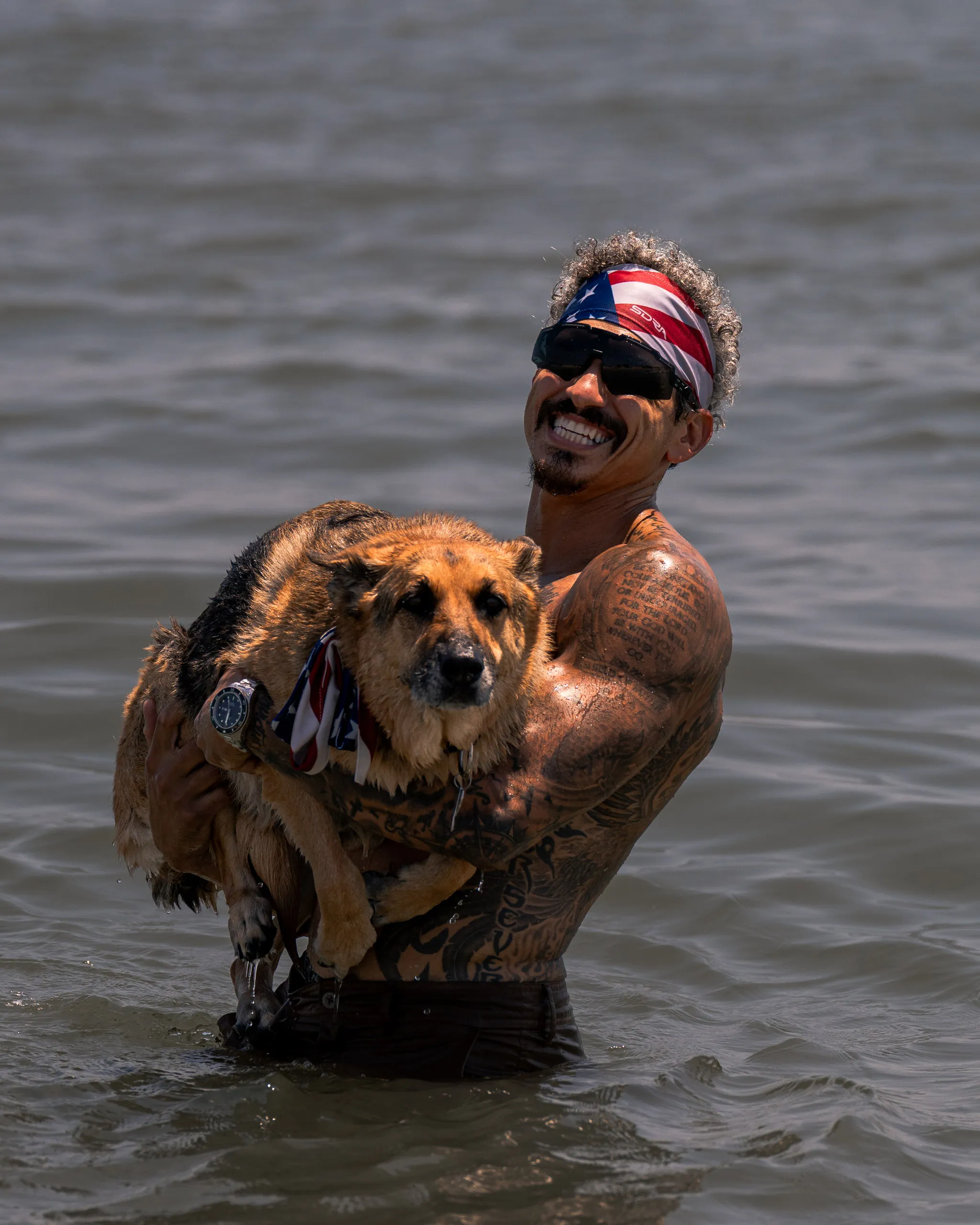 A man in a bandana carries his dog through chest-deep lake water