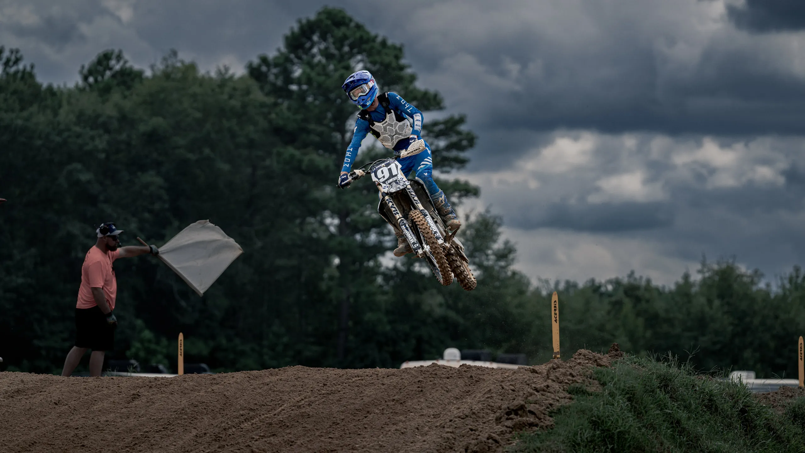 A rider floats a jump against heavy storm cloud while a trackside official waves a white flag