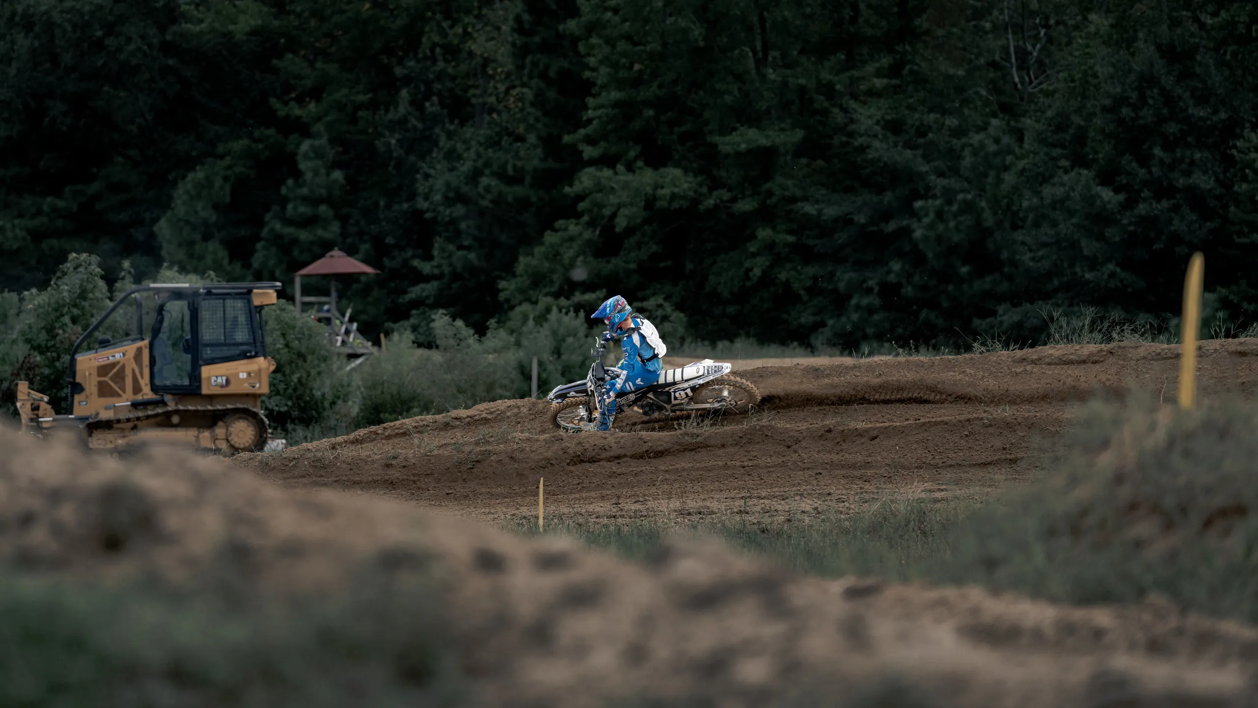 A lone rider works a far berm, seen across the track past a parked dozer and the timing tower in the trees