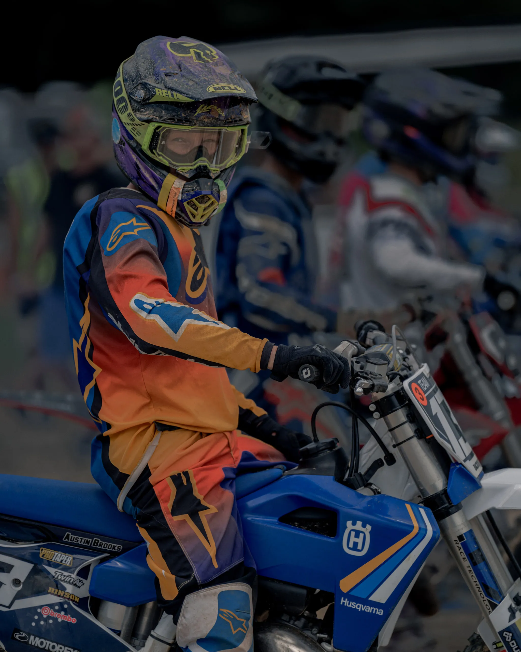 A young rider sits goggled up on a blue Husqvarna in the staging area, the rest of the gate blurred out behind