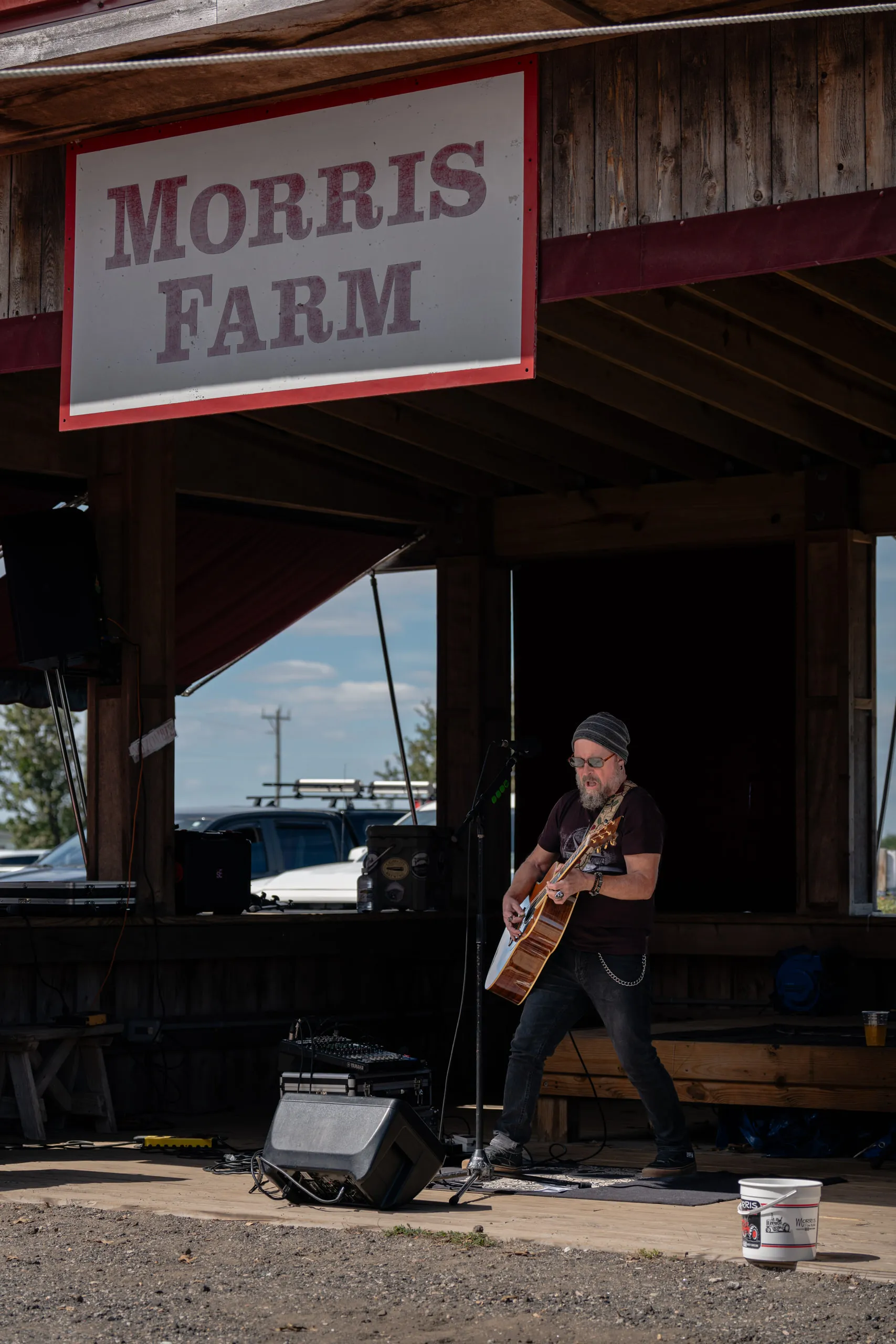 A guitarist plays beneath the Morris Farm sign on the covered stage, gear and a monitor set out alongside