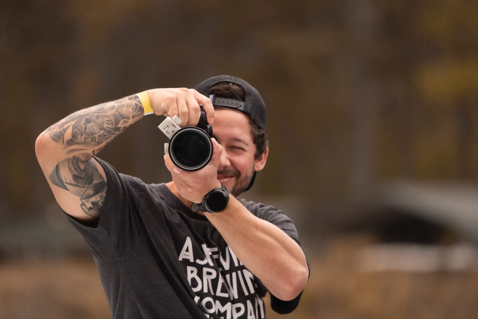Lou Cuevas raises a camera to his eye and grins over the top of it, autumn trees blurred behind him